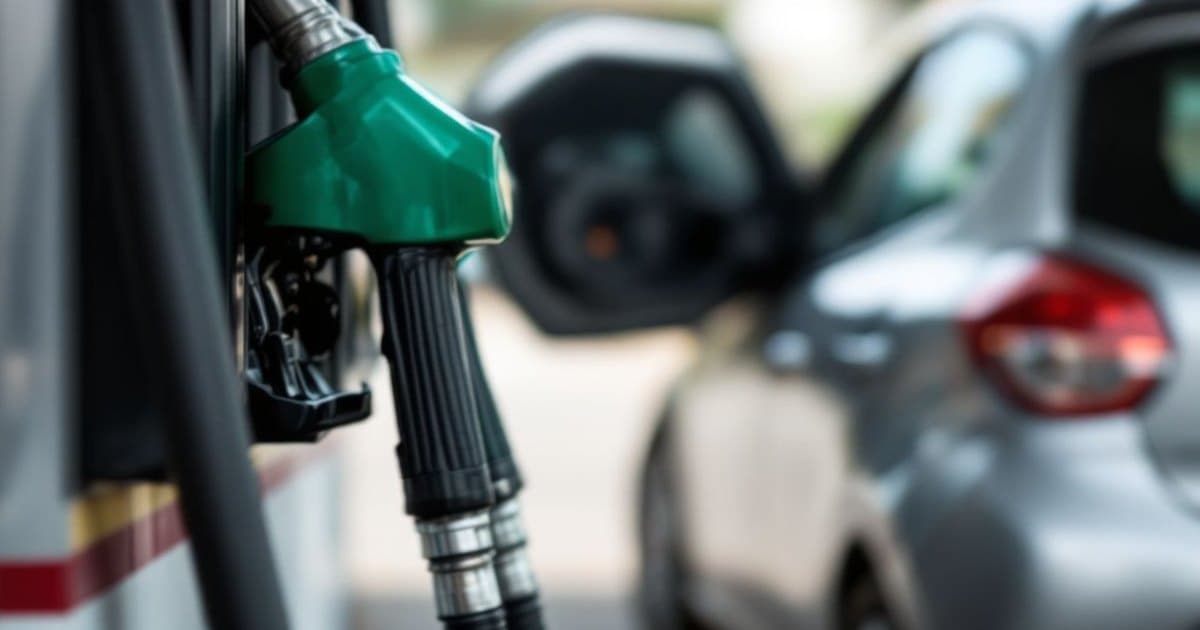 Bicos de abastecimento verde e preto pendurados numa bomba de posto, com um carro prata de porta aberta ao fundo, fora de foco