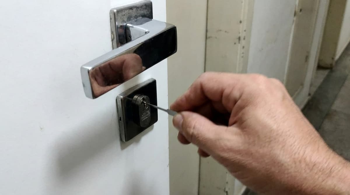 Mão do chaveiro com a ferramenta no miolo de uma porta residencial branca, vista do corredor