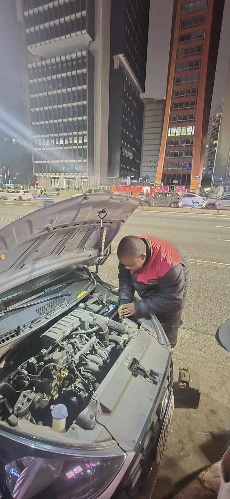 Técnico da Resgatou trocando a bateria de um carro à noite, com o capô aberto, na Avenida Paulista