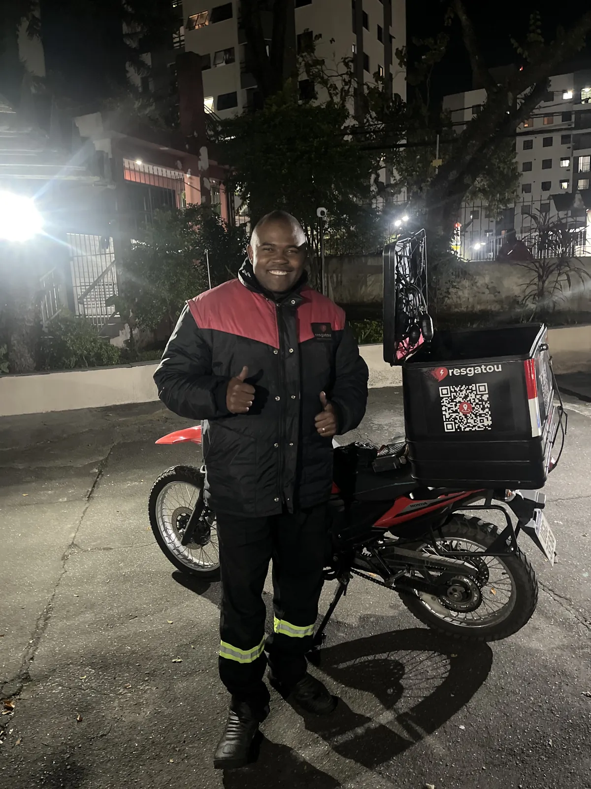 Técnico da Resgatou uniformizado ao lado da moto de atendimento com baú da empresa, à noite em rua de São Paulo